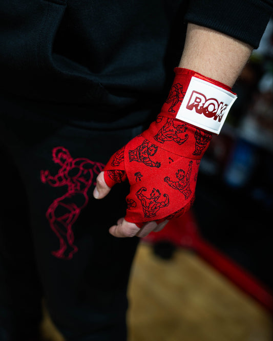 close-up of red anime boxing hand wraps with grappler illustrations and roki patch worn on fist, black athletic outfit background