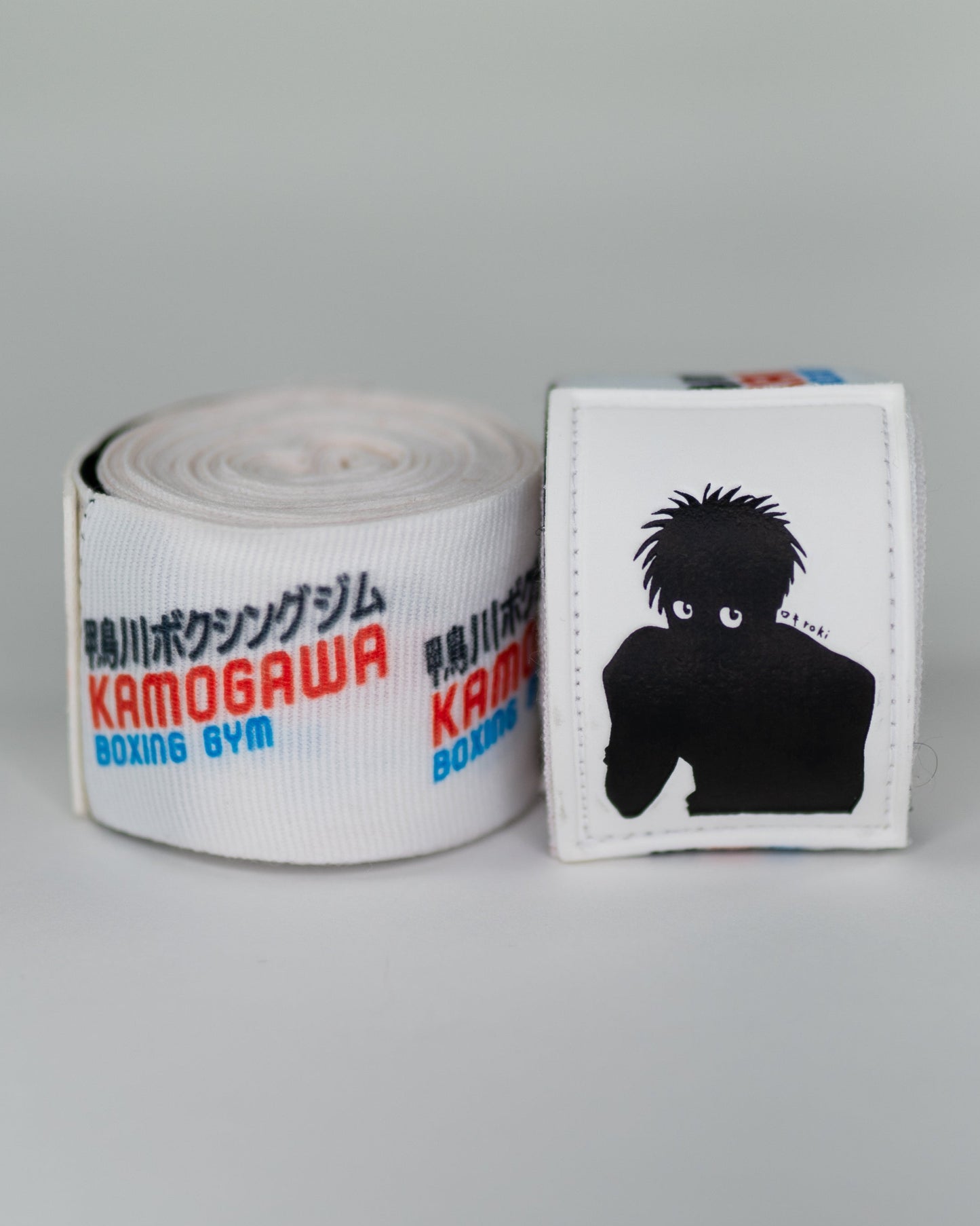 Close-up of anime boxing hand wraps featuring Kamogawa Boxing Gym logo and Ippo silhouette patch with rolled wrap