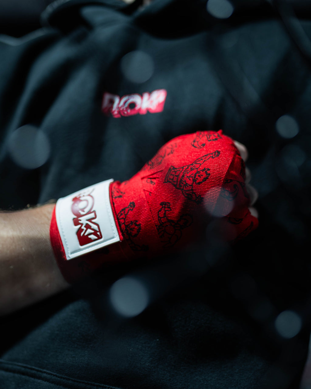 red anime-style boxing hand wraps with grappler figure print and roki patch, worn in closed fist against black hoodie background, with mma cage blur in front out of focus