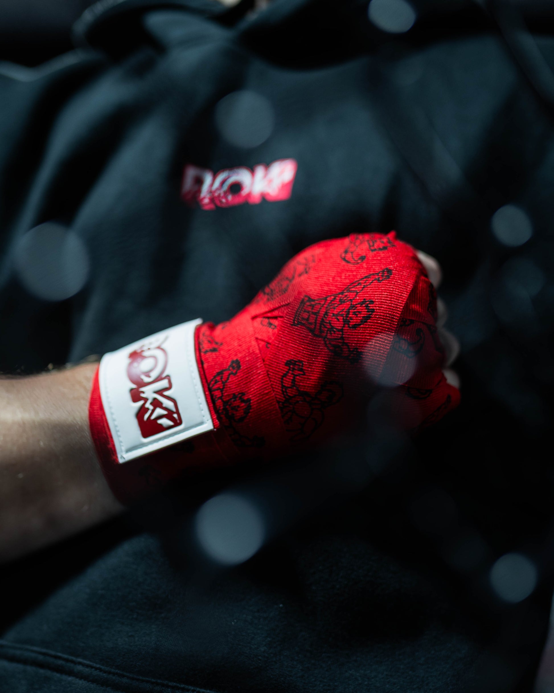 red anime-style boxing hand wraps with grappler figure print and roki patch, worn in closed fist against black hoodie background, with mma cage blur in front out of focus