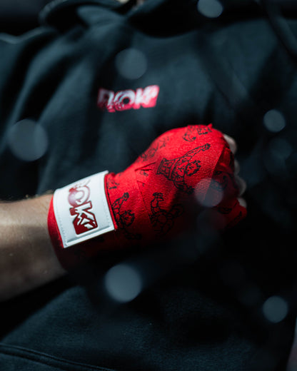 red anime-style boxing hand wraps with grappler figure print and roki patch, worn in closed fist against black hoodie background, with mma cage blur in front out of focus