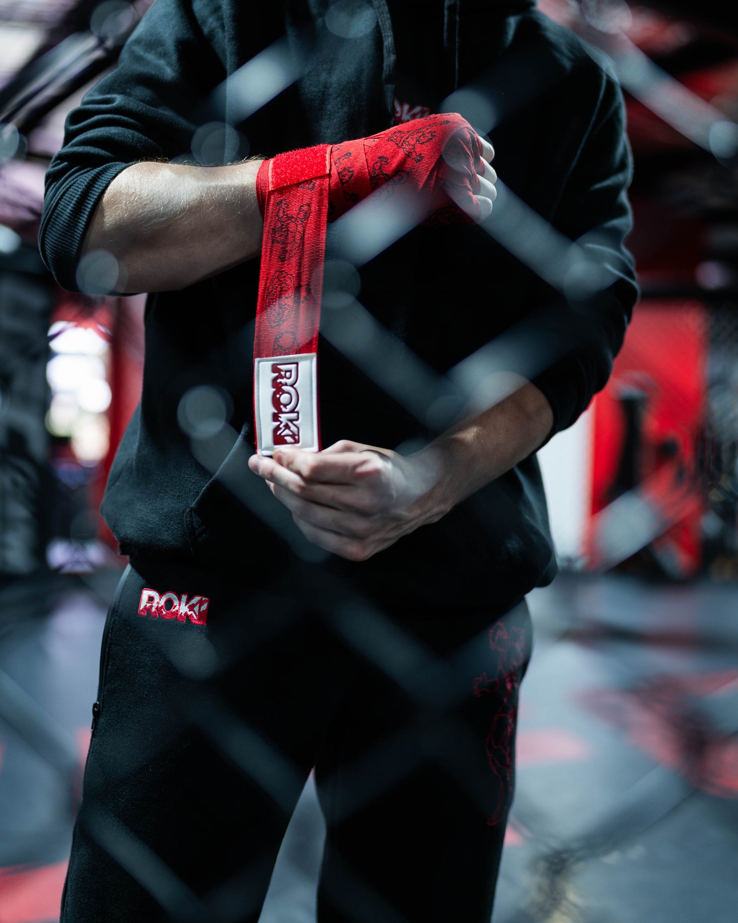 fighter tightening red anime-style boxing hand wraps with grappler graphics and roki patch inside mma cage environment