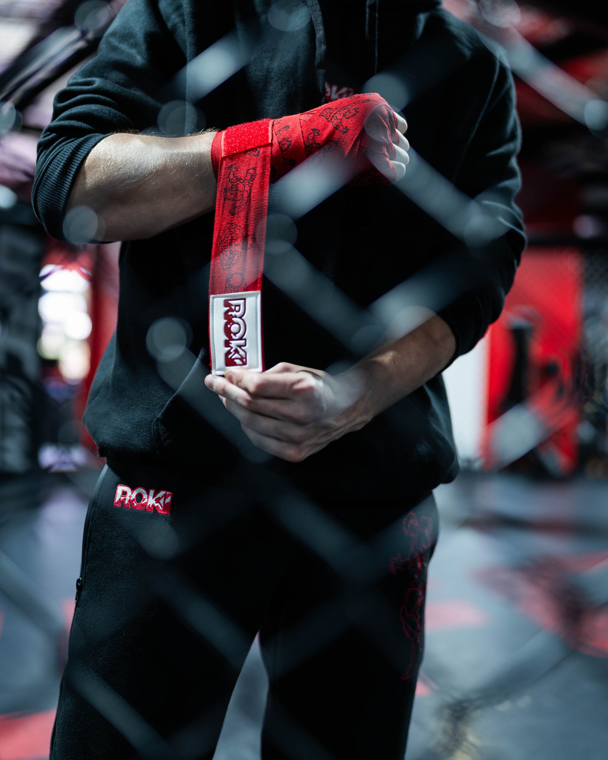 fighter tightening red anime-style boxing hand wraps with grappler graphics and roki patch inside mma cage environment