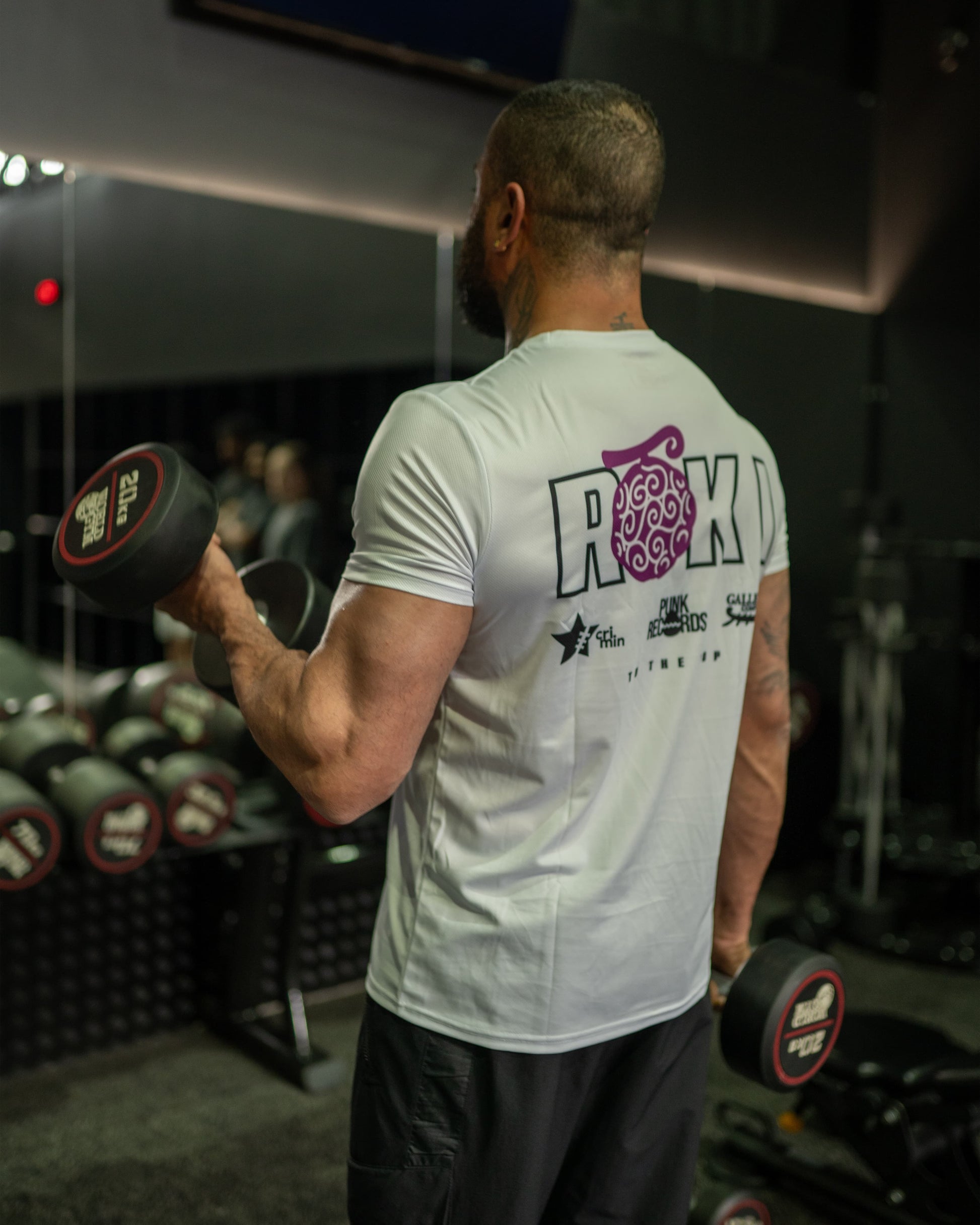 Back view of a model wearing a white Roki Liberation training shirt, lifting weights in a gym