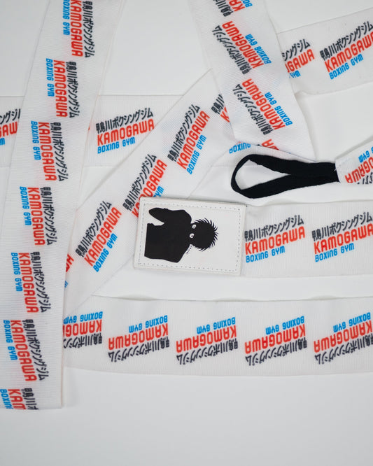 Flat lay of anime boxing hand wraps with Kamogawa Boxing Gym print and Ippo patch, showing thumb loop and repeating logo design cropped

